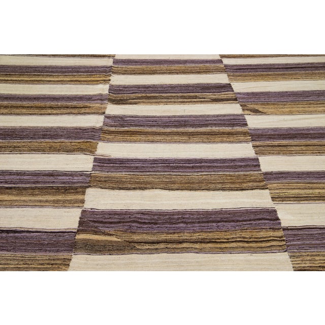 This contemporary kilim wool rug showcases a detailed and lively stripe design incorporating shades of beige, brown, and...