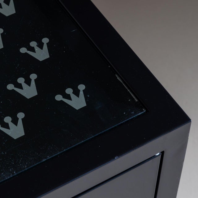 Black chest of drawers featuring four drawers resting on a square base. Its front is adorned with rows of stylized grey...