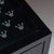 Black chest of drawers featuring four drawers resting on a square base. Its front is adorned with rows of stylized grey...