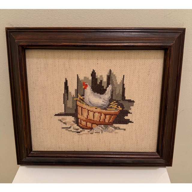 Charming vintage circa 1970s cross-stitch depicting a white and black chicken nestled in a woven basket, worked in a warm,...