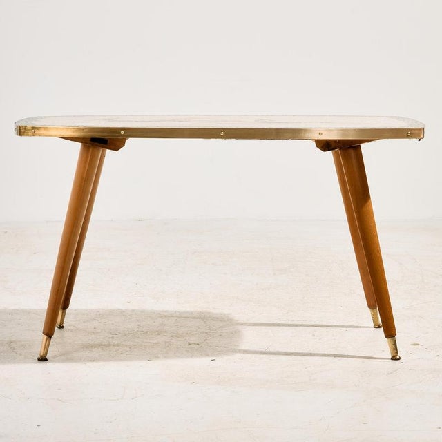 A mosaic-topped coffee table with organic patterns from the 1960s. The table’s edge and oak legs feature brass details and...