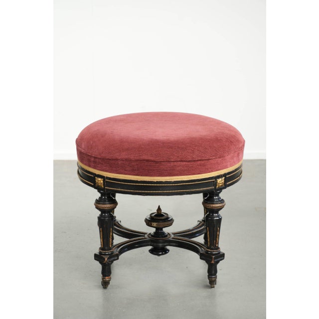 This antique English stool has been reupholstered in red fabric and features a black wooden base. The stool has casters...