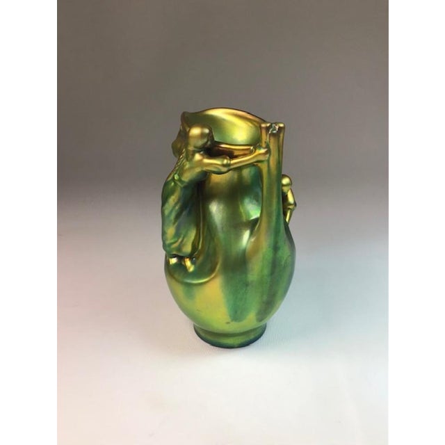 An iridescent Eosin green glaze pitcher depicting two reliefs of maidens gathering sticks. One maiden crouching, while the...