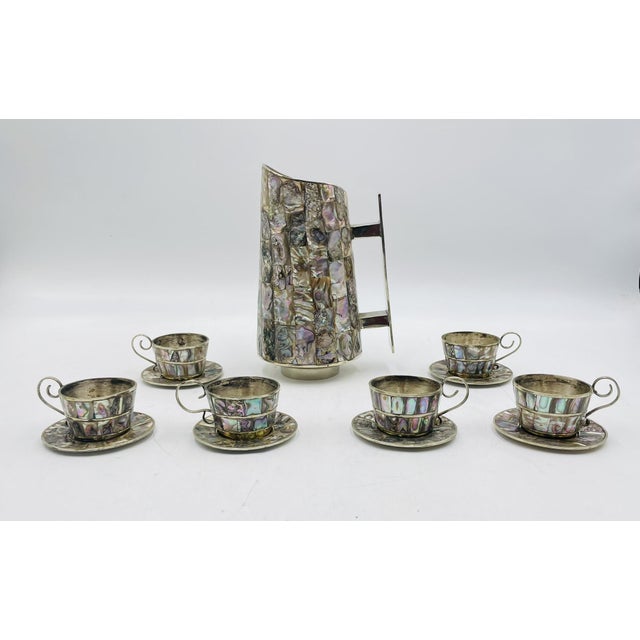 A striking and sculptural water pitcher with six matching cups and saucers, crafted in Mexico in the 1950s in alpaca...