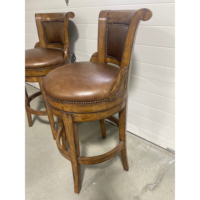 A pair of French Country style top grain leather upholstered swivel barstools of solid alder wood with antiqued brass nail...