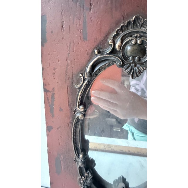 striking gothic-style wall mirror features an ornate, antiqued silver baroque frame mounted on a distressed red wooden...