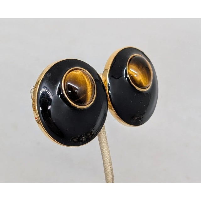 1970s goldtone round slightly domed black enamel with cabochon faux-tiger's eye at the top clip back earrings. Marked...