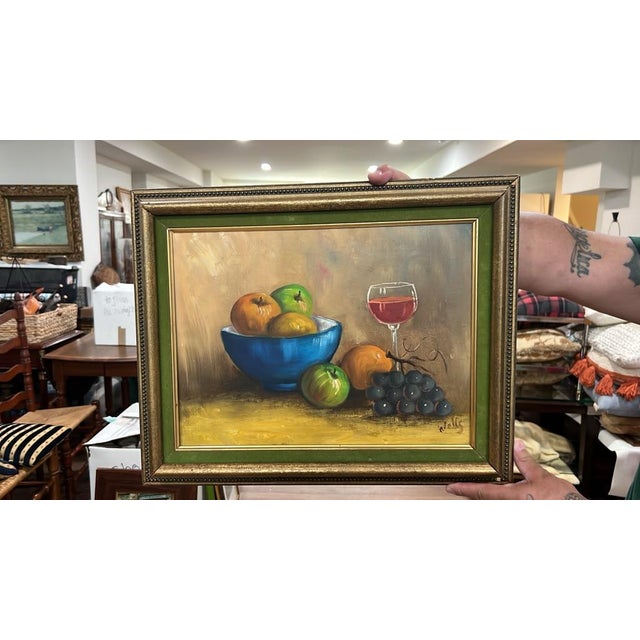 A classic still life oil painting depicting a vibrant blue bowl with fruit, apples, dark grapes, and a glass of red wine...