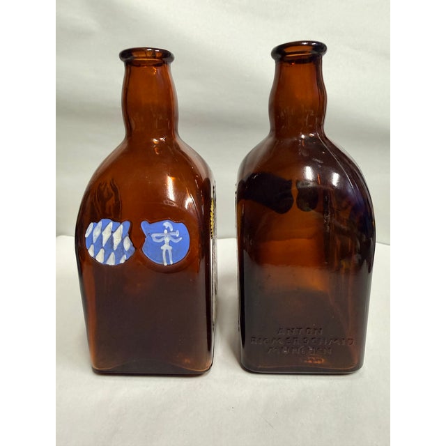 This vintage set includes two amber glass bottles featuring colorful hand-painted German designs on the front and sides....