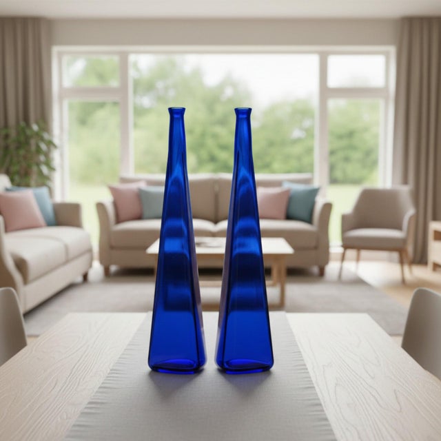 This matched pair of tall cobalt blue bottles reflects late 20th-century decorative glass production, emphasizing color...