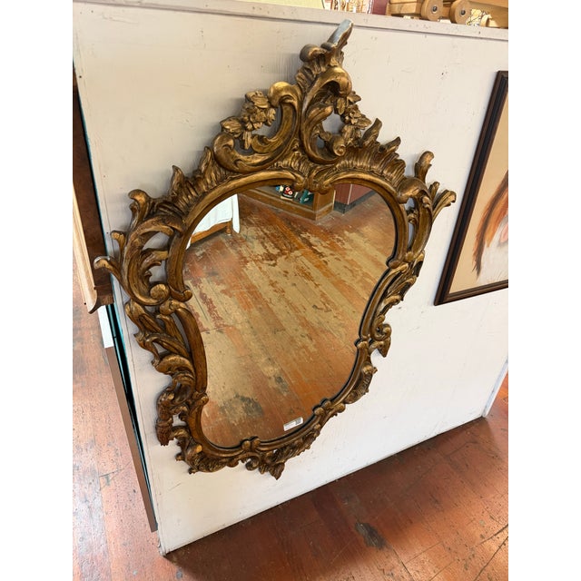 Elaborate gilt-finished wall mirror featuring an asymmetrical scrolling frame with floral and foliate detailing. It’s...