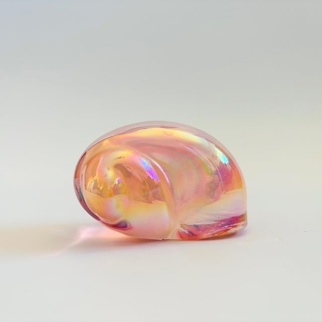 A vintage art glass paperweight in the shape of a shell. Iridescent pink color to the glass. Measures 3.5"W x 2"D x 2"H
