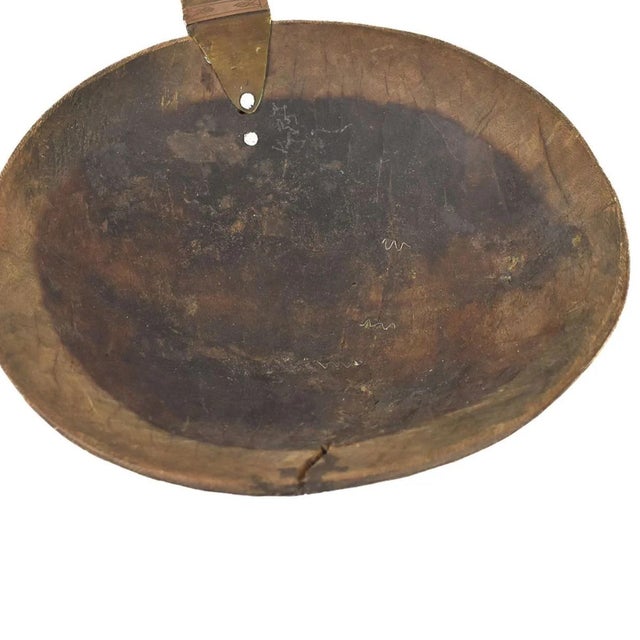 A large spoon or scoop made out of wood carved with a large bowl and a long handle with metal designs. This spoon is from...