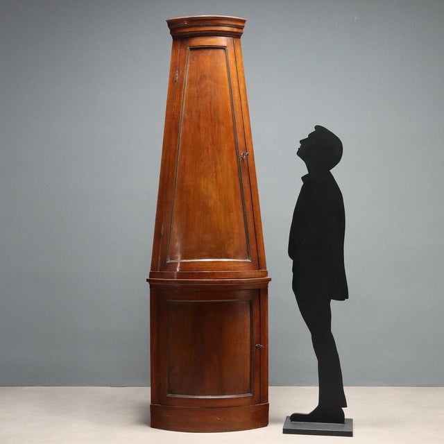 Walnut corner cabinet. The top has a truncated conical section and a paneled door, as does the base. Brass sheet moldings....
