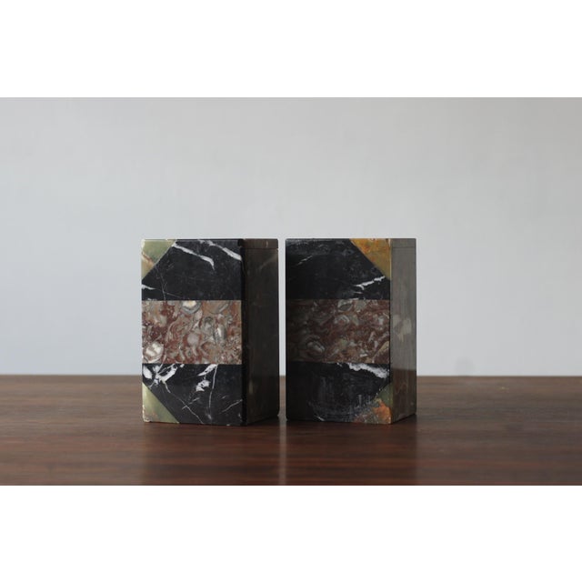 A pair of bookends made of natural, multicolored marble, featuring a compact, geometric design. The cuboid forms are...