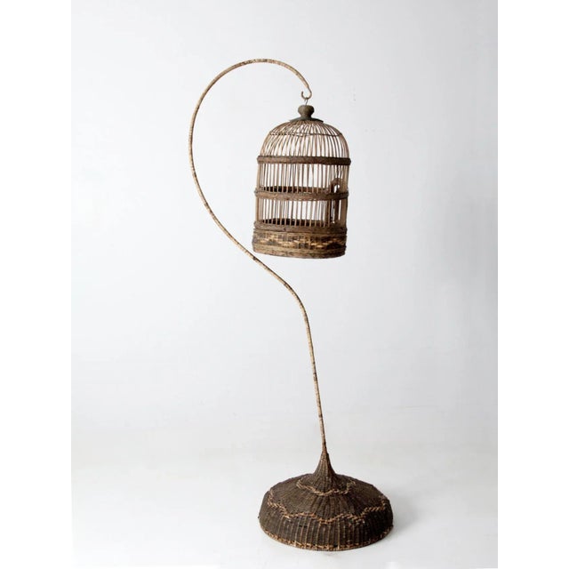 A vintage wicker bird cage with stand, featuring a hand-woven twig and rattan cage set atop a wood base with a metal...