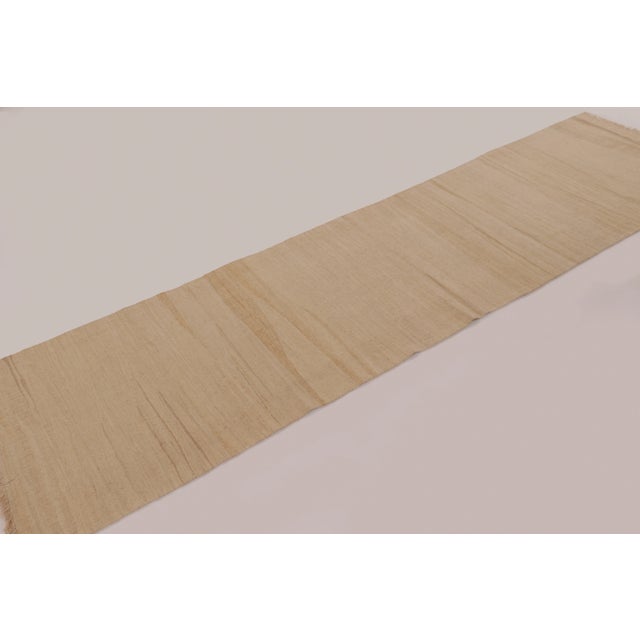 Handwoven in wool, this 3x10 contemporary Rez kilim presents a natural beige field enriched by subtle tonal variation and...