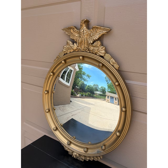 A Federal-style giltwood convex mirror crowned with a carved eagle, its wings outstretched above a laurel motif—an...