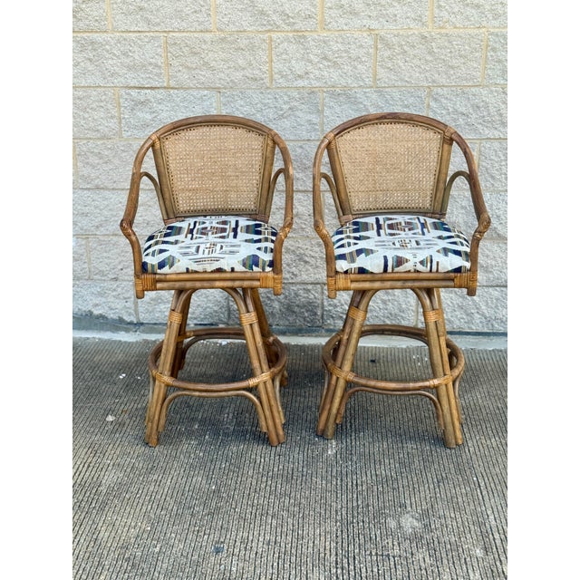 Pair of vintage rattan swivel bar stools, circa 1970s. Features woven cane backs, curved rattan arms, wrapped joint...