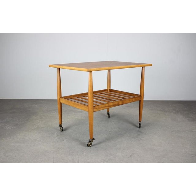 This Mid-Century Danish rolling table from the 1960s features a teak-veneered top, slatted lower storage shelf, and...