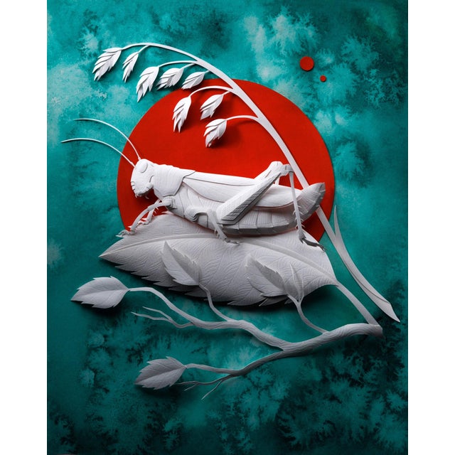 This hand-cut paper sculpture features a meticulously crafted grasshopper poised on a leaf, set against a vibrant red...