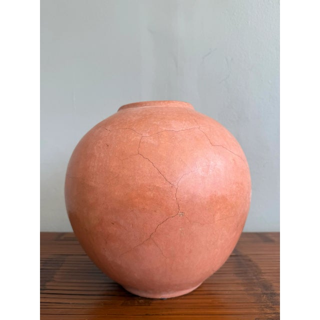 This rounded terracotta vessel bears a softly weathered surface, its warm, earthy hue interrupted by delicate hairline...