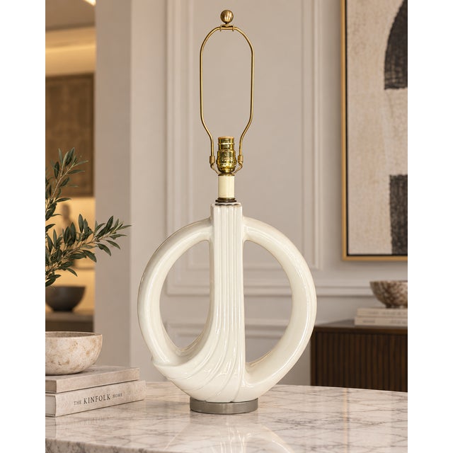 Table lamp with a glossy ivory-white ceramic body in an open-loop shape, with a central ribbed detail and brass-tone...