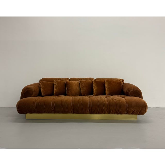 A dramatic sculptural sofa featuring oversized rolled arms, deep tufting, and a low-profile silhouette anchored by a...