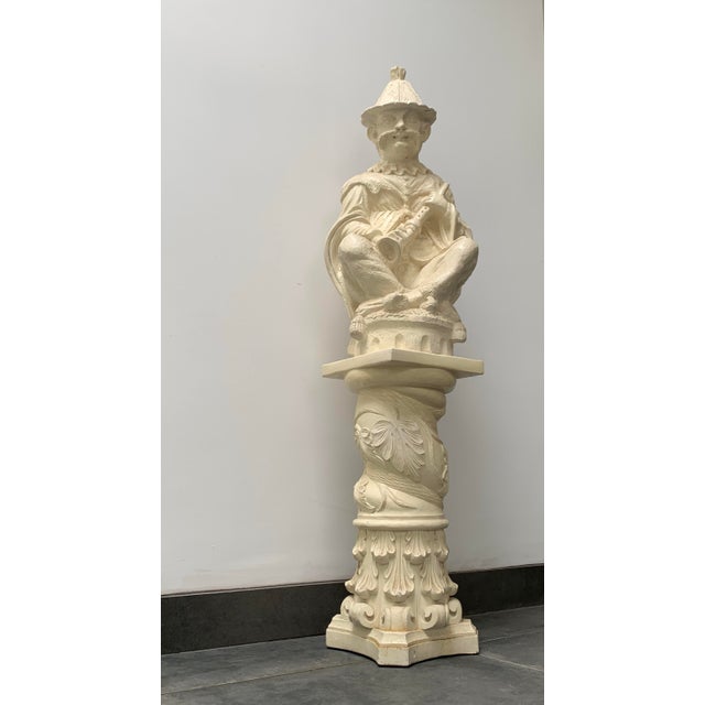An almost life size, white ceramic statue of a chinoiserie musician with flute on a pedestal. Seated crosslegged he is...