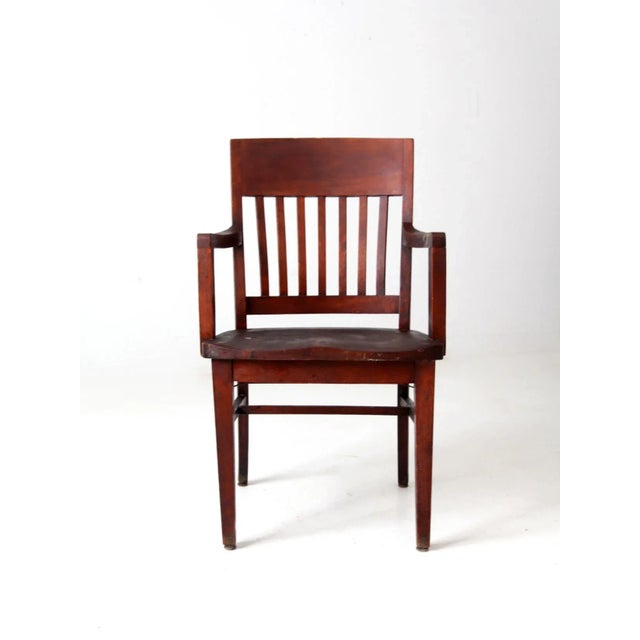 An early 20th-century Arts and Crafts-style wooden library armchair featuring a classic slat back, formed seat, and sturdy...