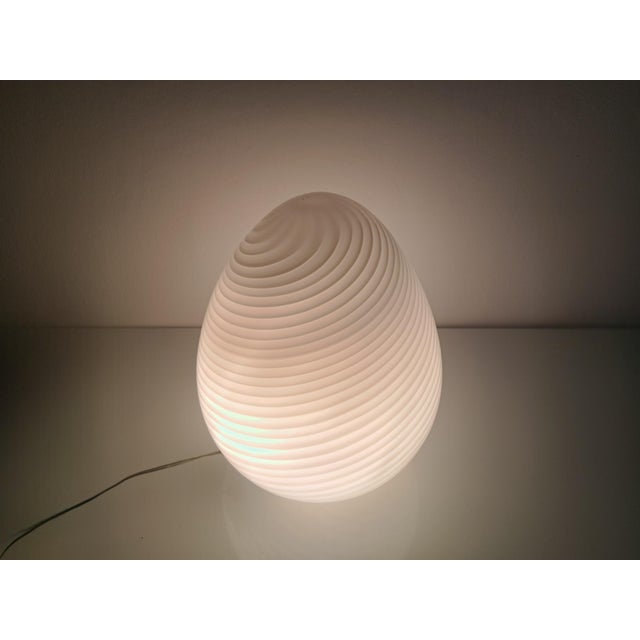 This Venetian egg-shaped Murano glass table lamp, crafted in the 1980s, is crafted using the Murano "rigadin ritorto"...