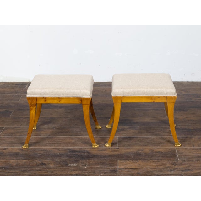 A pair of Austrian Biedermeier period walnut stools from the 19th century, with saber legs, carved giltwood lion paw feet...