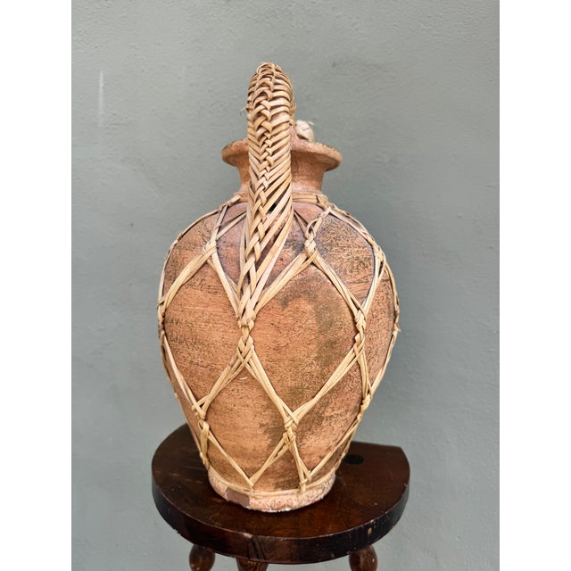 A large terracotta amphora encased in a precise hand-woven rattan lattice from shoulder to base. Organic tension of...