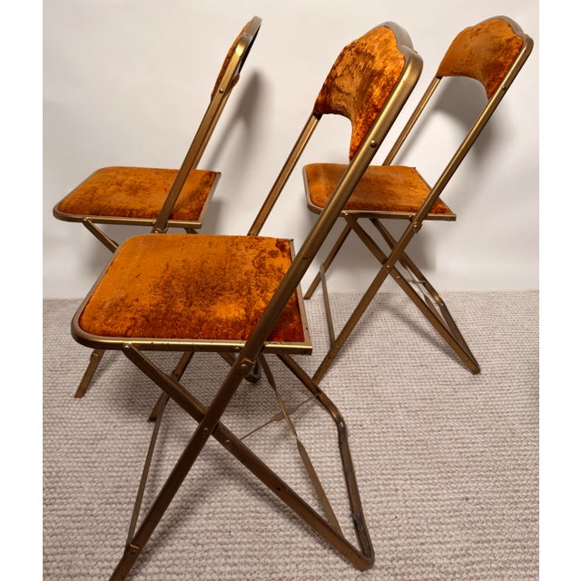 Vintage folding banquet chairs by Fritz & Co., featuring brass-tone metal frames and rust velvet upholstery. These chairs...