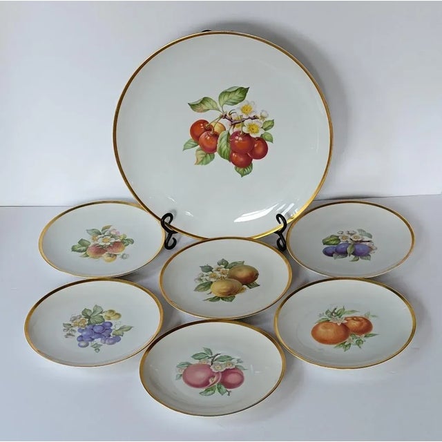 Circa 1960, six porcelain 8" luncheon or dessert plates and a 13.25" round serving platter in a fruit pattern. In near...