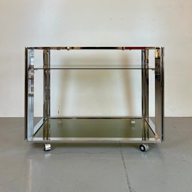 Elegant Space Age bar cart featuring a geometric chrome-plated metal frame and three shelves in olive-green smoked glass....