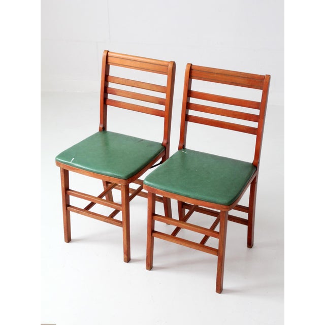 A pair of vintage 1950s folding chairs featuring warm reddish wood frames, slatted ladder backs, and green upholstered...