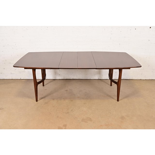 A sleek and stylish Mid-Century Modern sculpted walnut boat-shaped extension dining table Attributed to Lenoir Furniture...