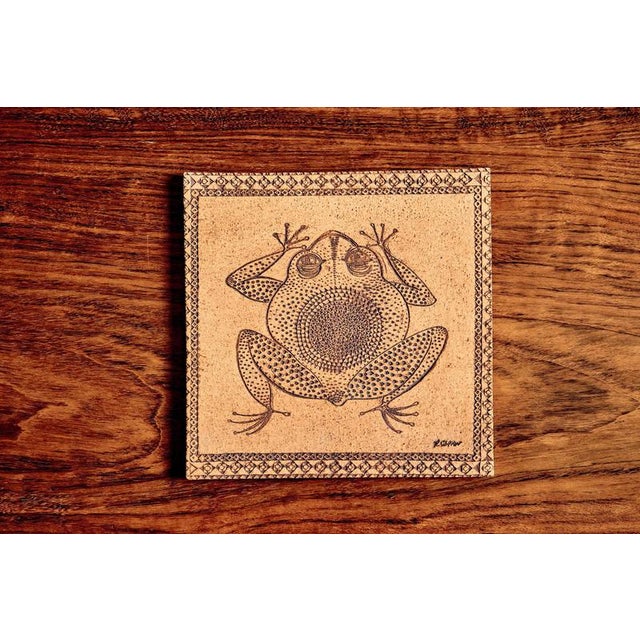 This square ceramic tile depicts a stylized frog rendered in incised line and dotted relief, framed by a geometric border....