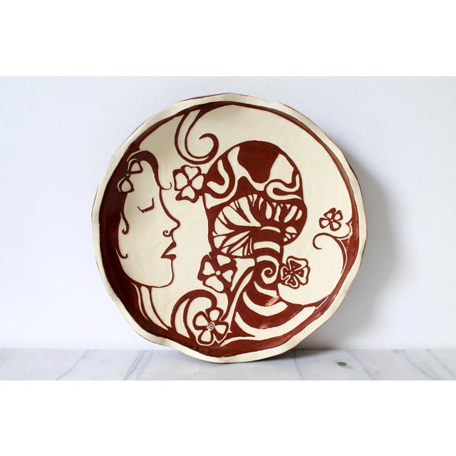 Whimsical brown and cream colored pottery plate with a female, mushroom and floral motif. Signed by artist. Minor age wear.