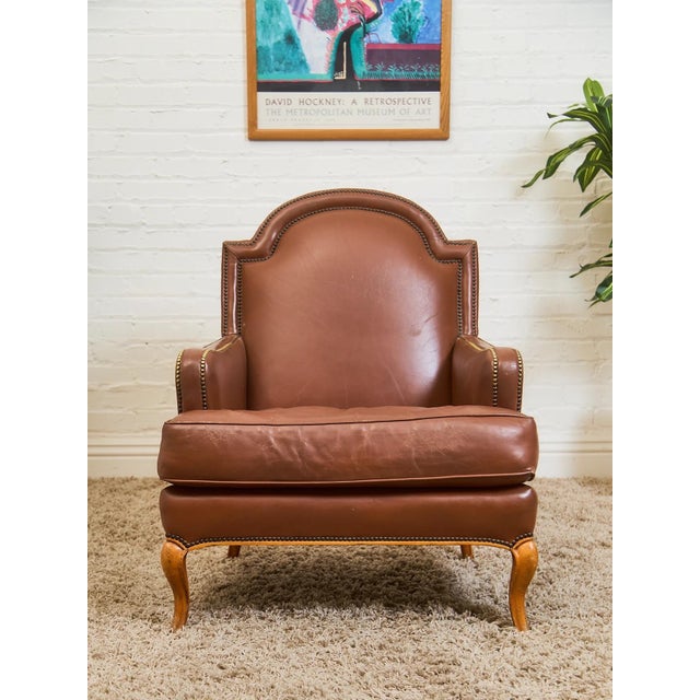 A stately leather bergère-style armchair featuring a gracefully arched back, rolled arms, and hand-applied brass nailhead...