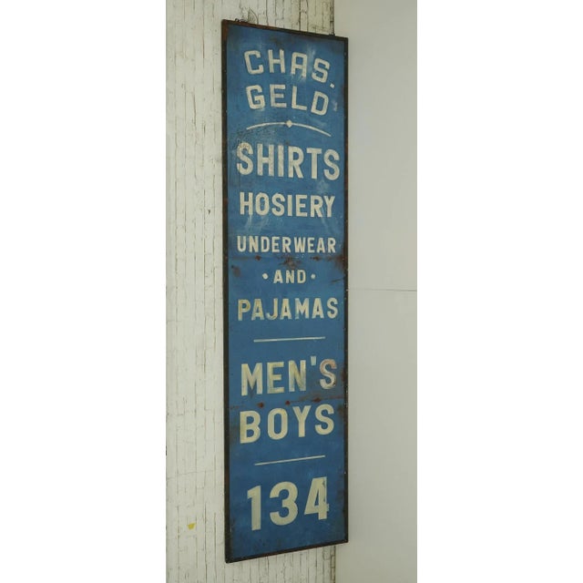 Reclaimed movie set sign made of aluminum in blue, white, and black colors. The sign reads "Chas. Geld, Shirts Hosiery,...
