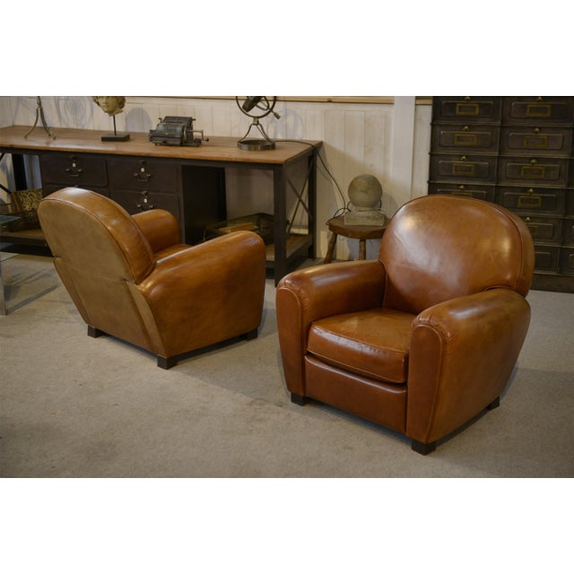 Pair of cognac-colored leather club chairs, offering enveloping comfort and generous proportions. with their rounded...