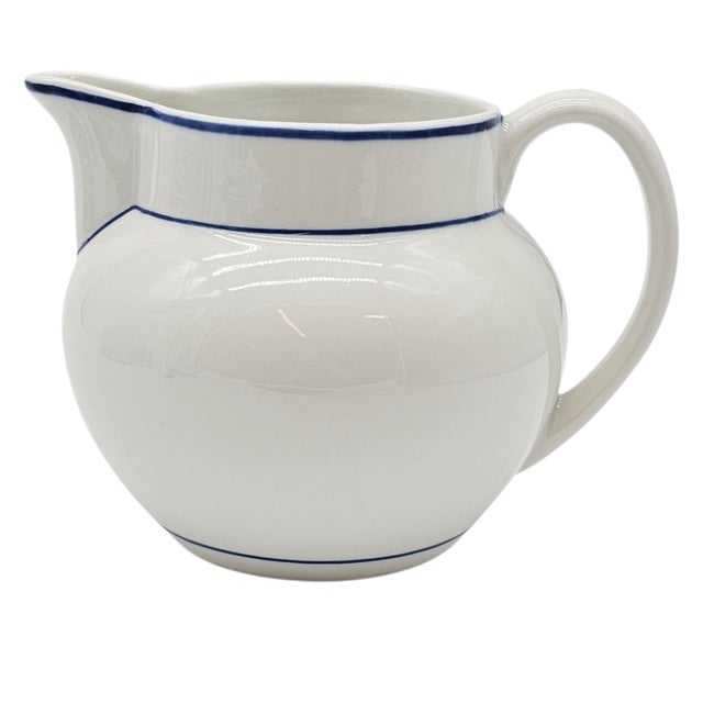 A pitcher that whispers of sunlit mornings and whitewashed terraces, its creamy surface adorned with a delicate blue rim –...