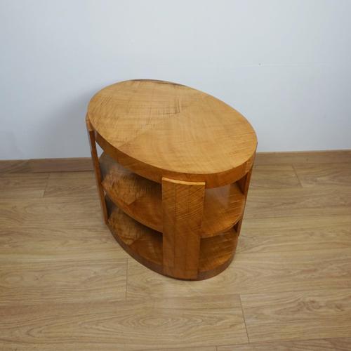 An Art Deco oval side table. Satin birch venereed throughout with two open shelves. Dimensions: H 56cm W 67.5cm D 47cm...