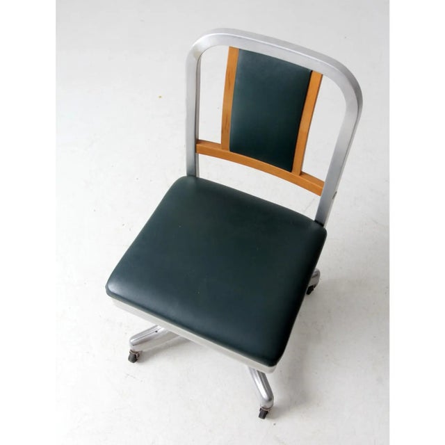 A classic office design featuring an aluminum base with casters, a curved wood back, and a green vinyl upholstered seat....