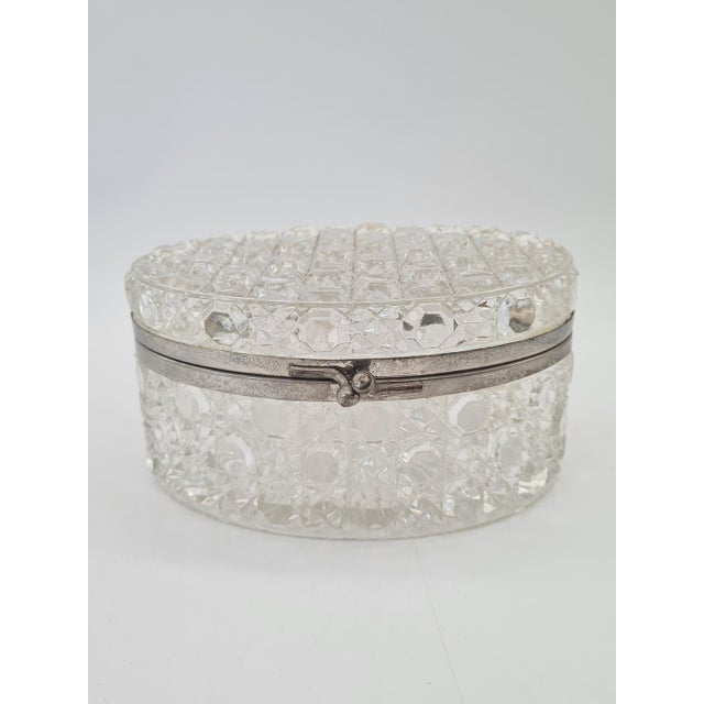 An elegant, oval-shaped box made of richly faceted pressed glass, with a decorative hinged lid and a metal frame. The...