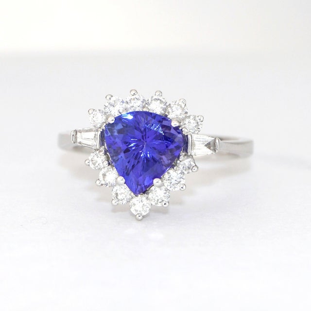 This elegant ring is crafted in 18K white gold. It features a striking trillion-cut tanzanite, surrounded by a halo of...