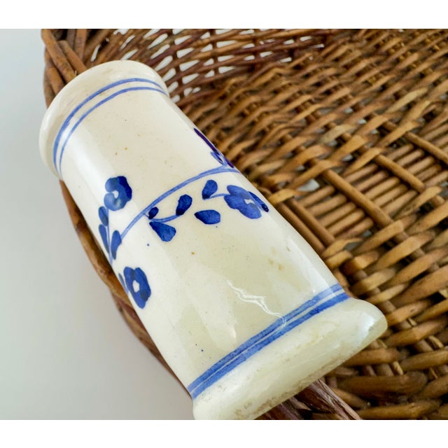 A distinctive vintage woven wicker basket featuring a broad oval form and a pair of blue-and-white porcelain handles. The...
