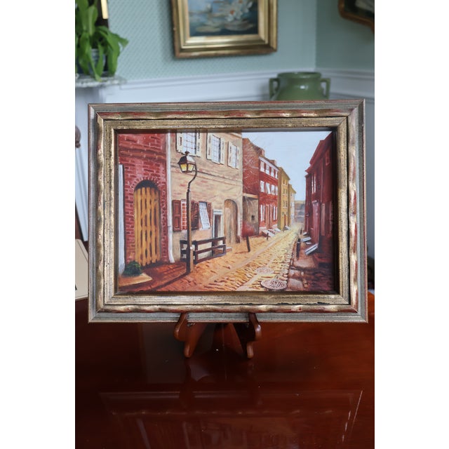 A signed original oil painting by Indiana artist Roger Merkel (1949–2016), depicting a narrow cobblestone street lined...